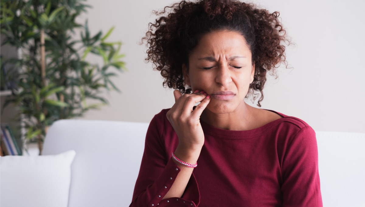 Woman sitting on a white couch with her eyes closed, fingers on her jaw and wincing in pian