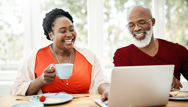Older attractive couple looking at a laptop and smiling while eating a meal.