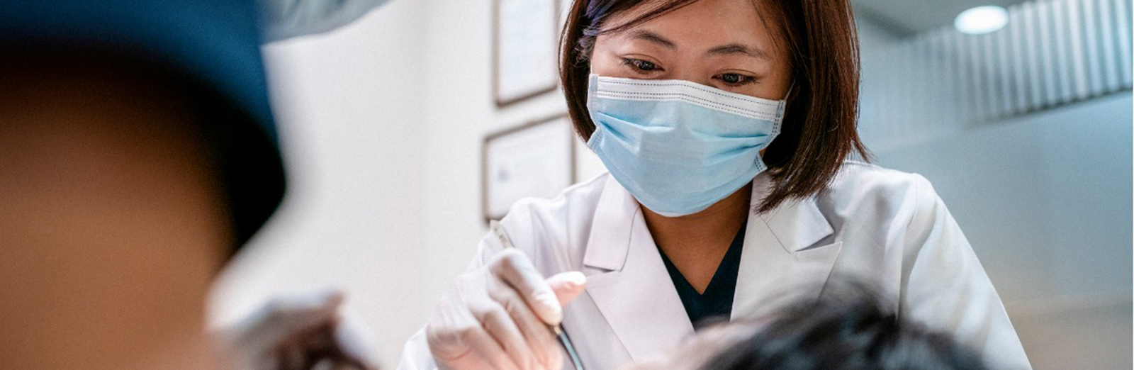 Dentist and her assistant wearing blue face masks examine a patient 1600x522