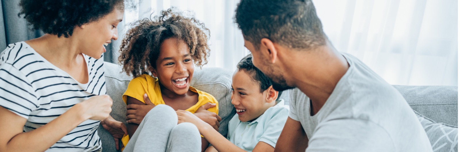 1600x529 Young family sitting on a couch at home talking and laughing. Mom dad and little brother are all looking at a little girl with a huge smile who is looking back at her dad.