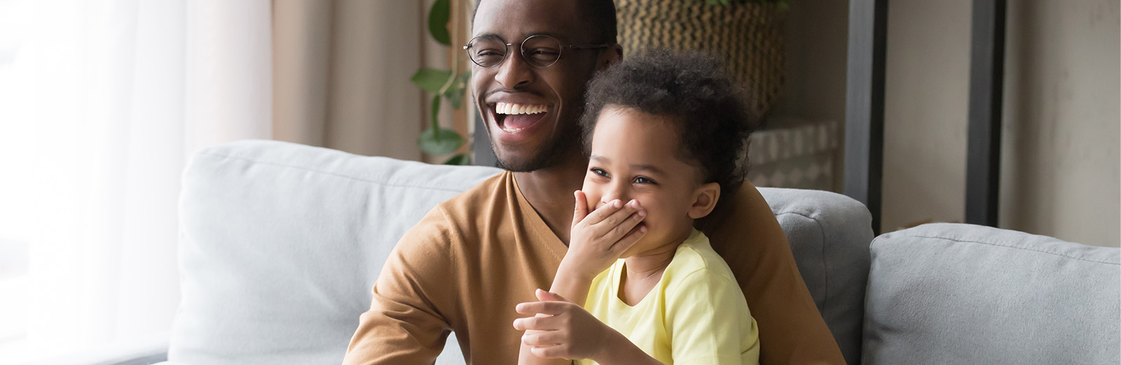 A young dad with a cute preschool aged child on his lap sitting on a couch. They are eating a big bowl of popcorn and laughing looking presumably toward a television. 