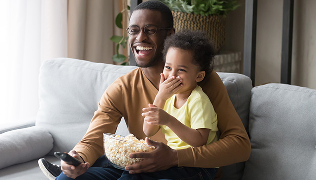 Thumbnail of a young dad with a cute preschool aged child on his lap sitting on a couch. They are eating a big bowl of popcorn and laughing looking presumably toward a television. 