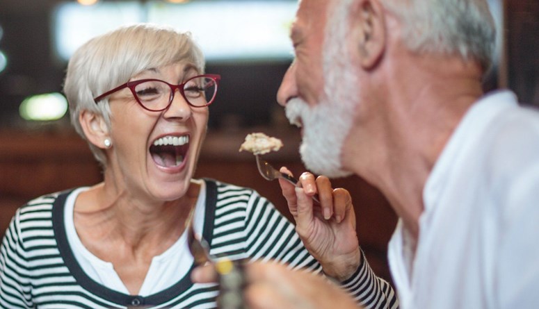 older woman in red framed glasses feeding her male partner a bite of food in a restaurant small