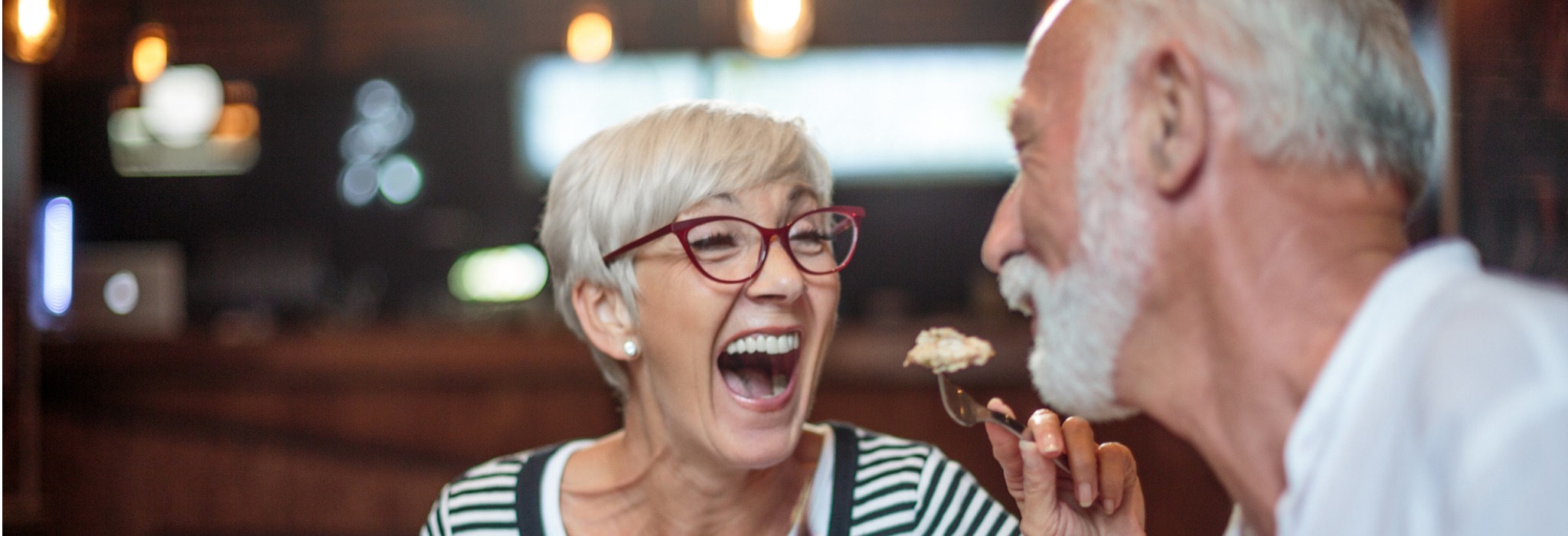 older woman wearing red framed glasses feeding her male partner a bite of food in a restaurant 2048x800