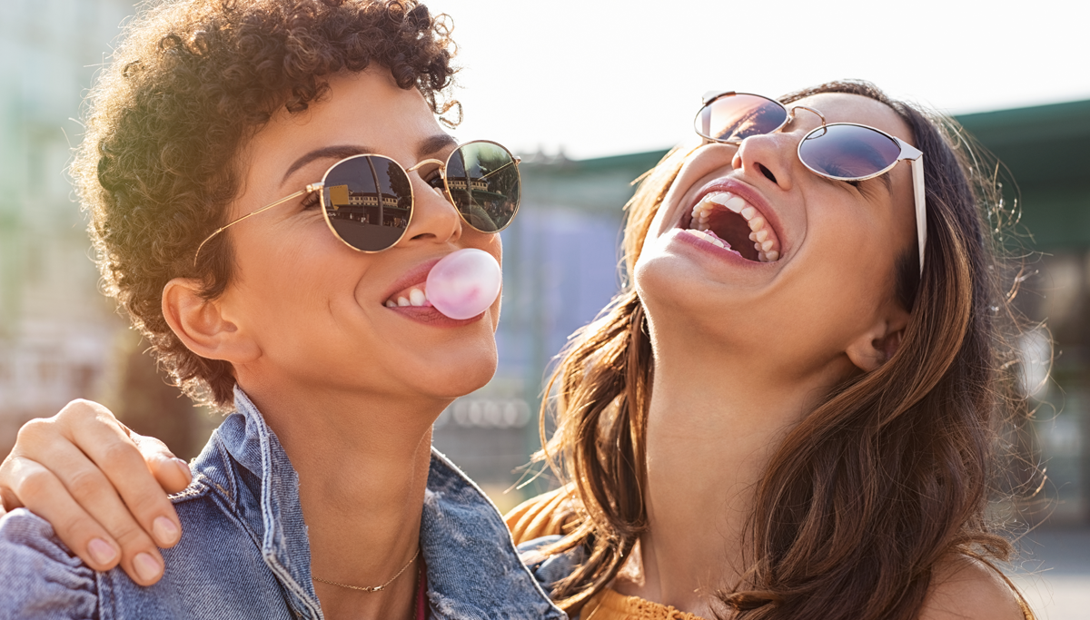 Two women with sunglasses outside in the sun smiling and laughing